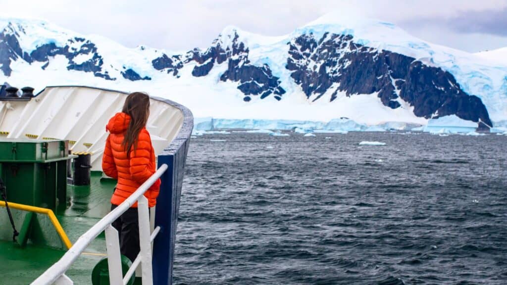 A person in an orange jacket stands on a ship’s deck, looking out at icy mountains and ocean near Antarctica.