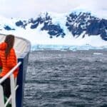 A person in an orange jacket stands on a ship’s deck, looking out at icy mountains and ocean near Antarctica.