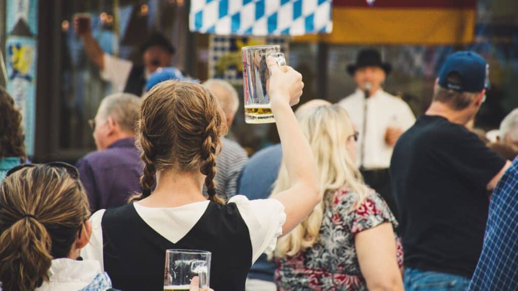A crowd of people, some holding beer mugs, attends an outdoor event reminiscent of Oktoberfests across America, with a musician performing in the background.