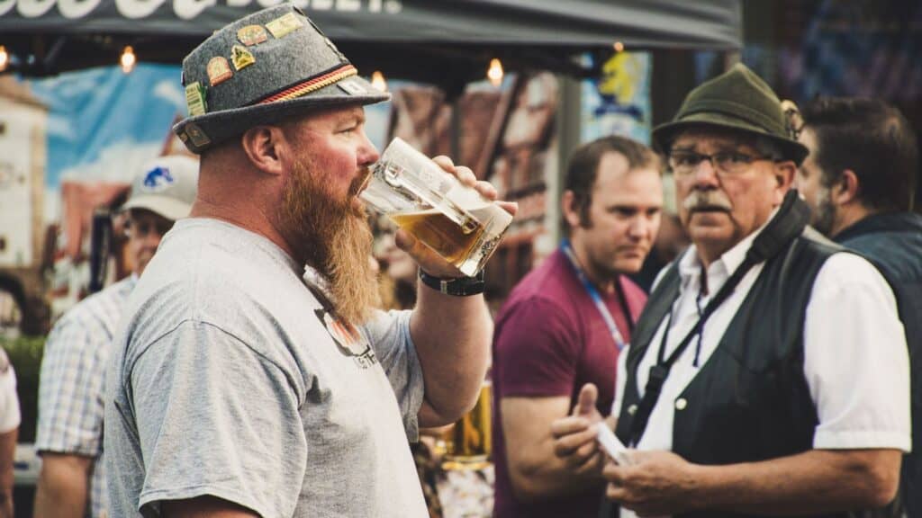 A man with a beard drinks beer from a glass while others talk in the background at an outdoor event, capturing the lively spirit found at Oktoberfests across America.