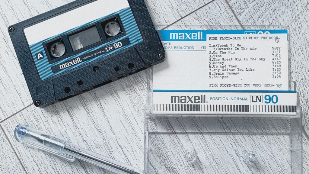 Maxell audio cassette tape labeled “Pink Floyd-Dark Side of the Moon” with handwritten tracklist—capturing classic mixtape making teenager rituals—clear plastic case, and a pen placed on a light wooden surface.
