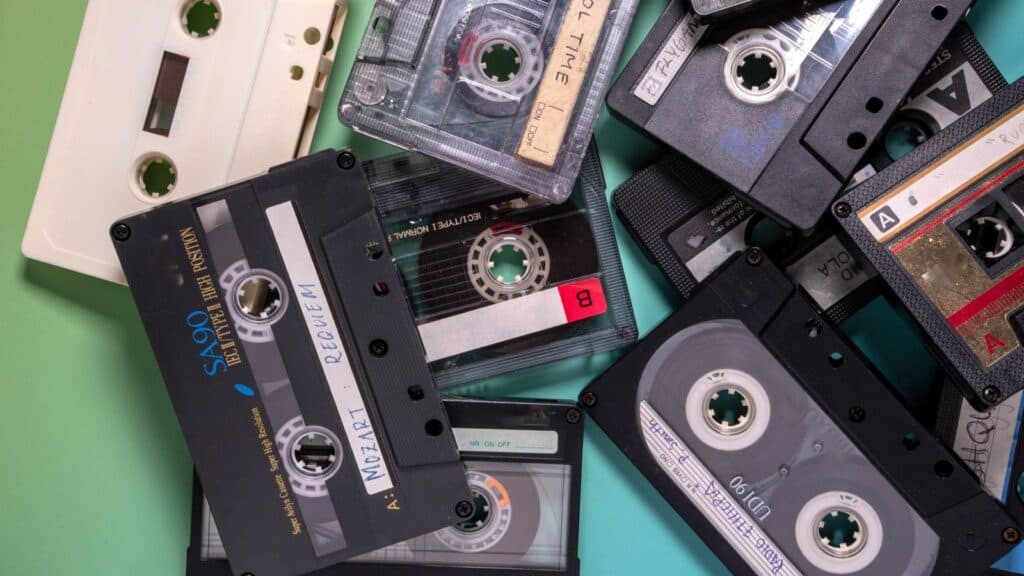 A scattered pile of various audio cassette tapes with handwritten and printed labels sits on a green surface, evoking memories of taped shows on VHS and the analog era.