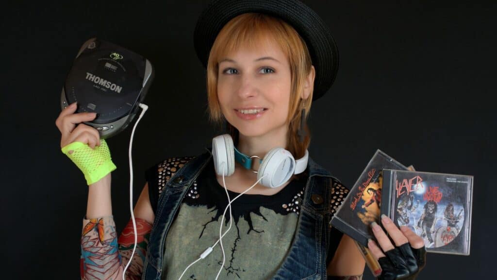 A person in a hat and headphones holds a portable CD player and three rock music CDs, standing against a dark background—a modern-day Walkman owner remembers listening to music in their own unique way.