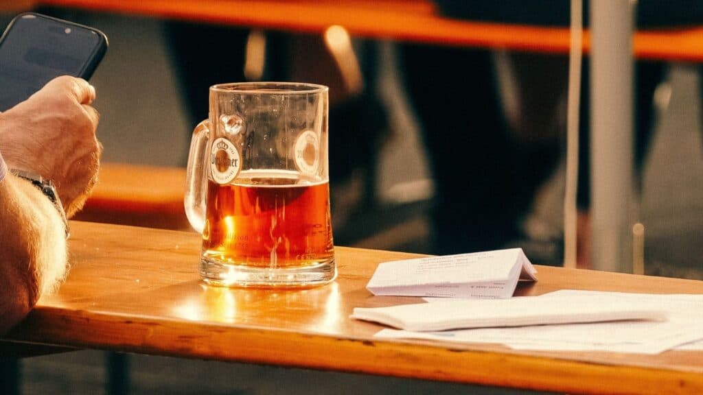 A person holding a smartphone sits at a wooden table with a half-full beer mug and some papers in sunlight, perhaps reading the fine print that explains what travel insurance does not cover.