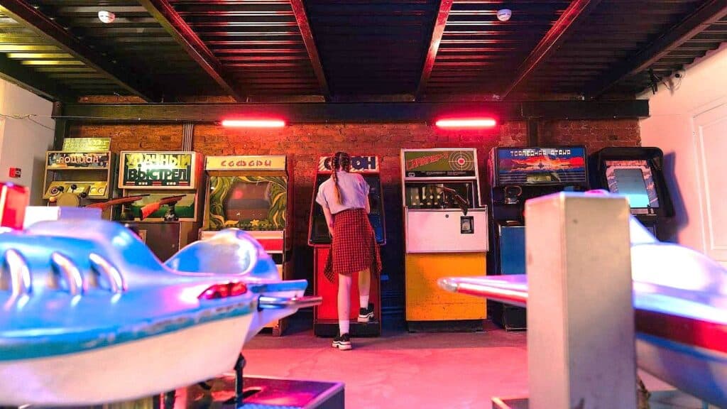 A person stands facing old 80s arcade games in a dimly lit retro arcade, with two blue spaceship game rides in the foreground.
