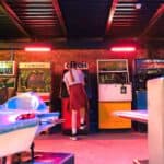 A person stands facing old 80s arcade games in a dimly lit retro arcade, with two blue spaceship game rides in the foreground.