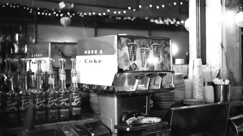 Black-and-white photo of a drink station with a soda fountain, Torani syrup bottles, stacked cups, and straws on a counter, evoking classic drugstore soda fountain traditions, with string lights visible in the background.
