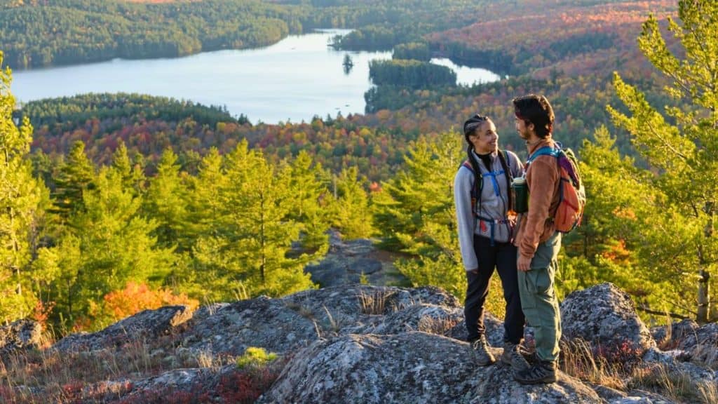Two hikers stand on a rocky outcrop overlooking a lake and forested hills with vibrant autumn foliage under a clear sky, capturing the beauty of the Adirondacks during peak leaf peeping season.