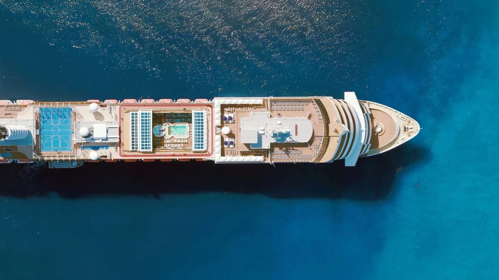 Aerial view of a large cruise ship with a pool, sports court, and deck areas, floating in clear blue water.