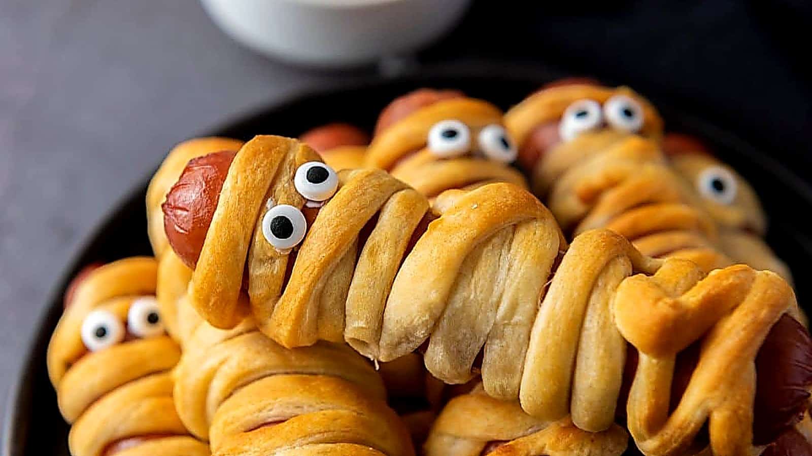 Hot dogs wrapped in crescent dough to resemble mummies, topped with candy eyes, arranged on a black plate with a small bowl of dipping sauce in the background.