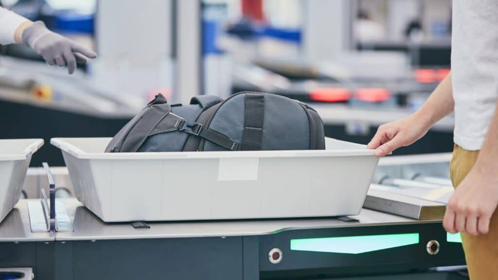 A person places a backpack in a plastic bin at an airport security checkpoint, preparing for TSA liquid screening at US airports, while another person gestures nearby.