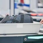 A person places a backpack in a plastic bin at an airport security checkpoint, preparing for TSA liquid screening at US airports, while another person gestures nearby.
