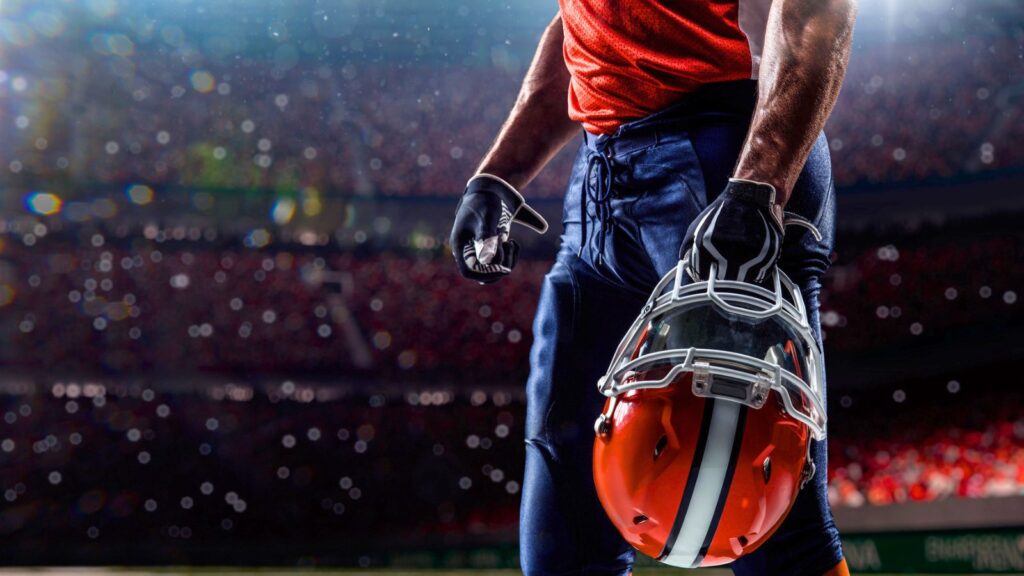 Close-up of an American football player in uniform holding a helmet on the field, with a stadium and crowd blurred in the background.
