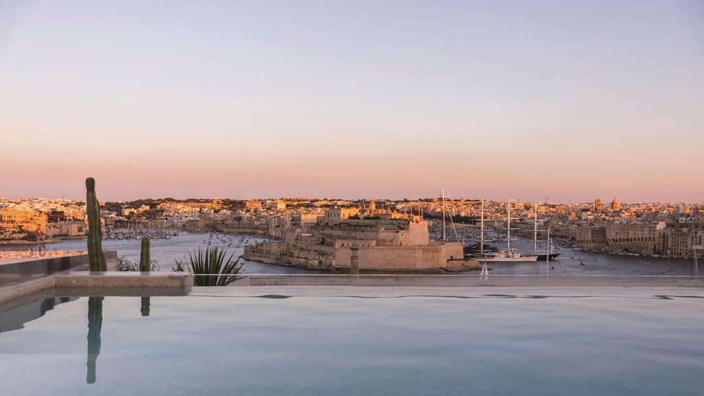 A sunset view of a harbor and coastal town with a stone fortress, seen from the infinity pool at Iniala Valletta.