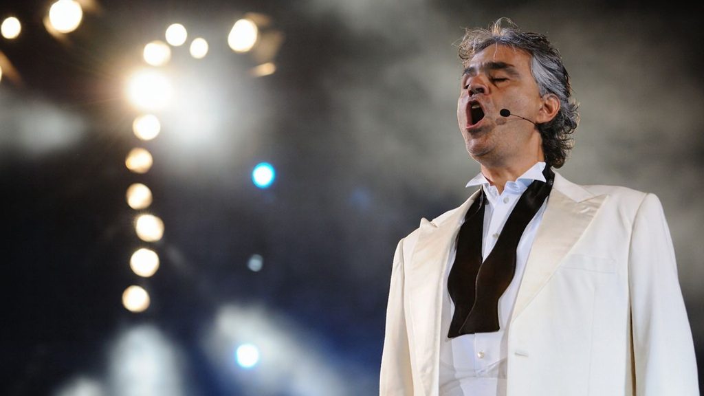 A man in a white tuxedo sings passionately on stage under bright lights, with a dramatic dark and smoky background.
