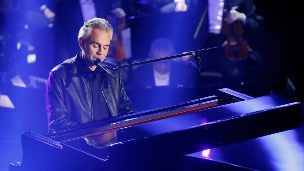 A man in a leather jacket plays a grand piano and sings into a microphone on stage, with an orchestra seated in the background.