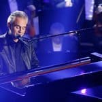 A man in a leather jacket plays a grand piano and sings into a microphone on stage, with an orchestra seated in the background.