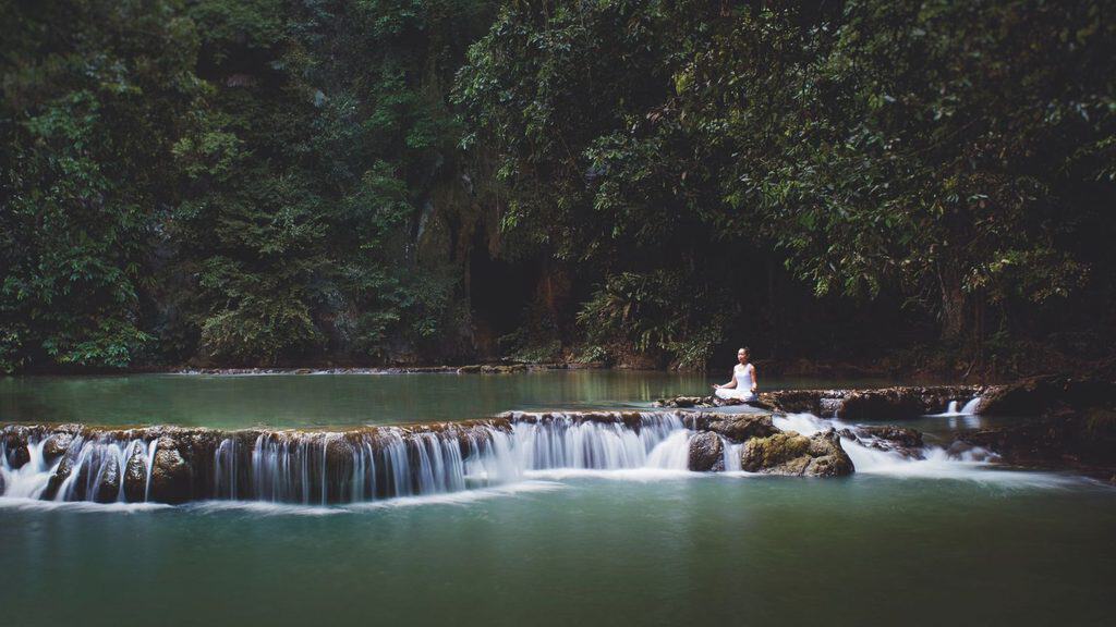 A person sits cross-legged meditating on a rocky ledge above a small waterfall in a forested area beside a calm green pool, reminiscent of the tranquil landscapes found in Phulay Bay, Krabi.