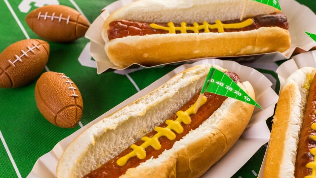 Three hot dogs with mustard laces resembling footballs sit on a football-themed tablecloth next to small toy footballs, offering a playful nod to discontinued NFL stadium foods that once left fans cheering for more.