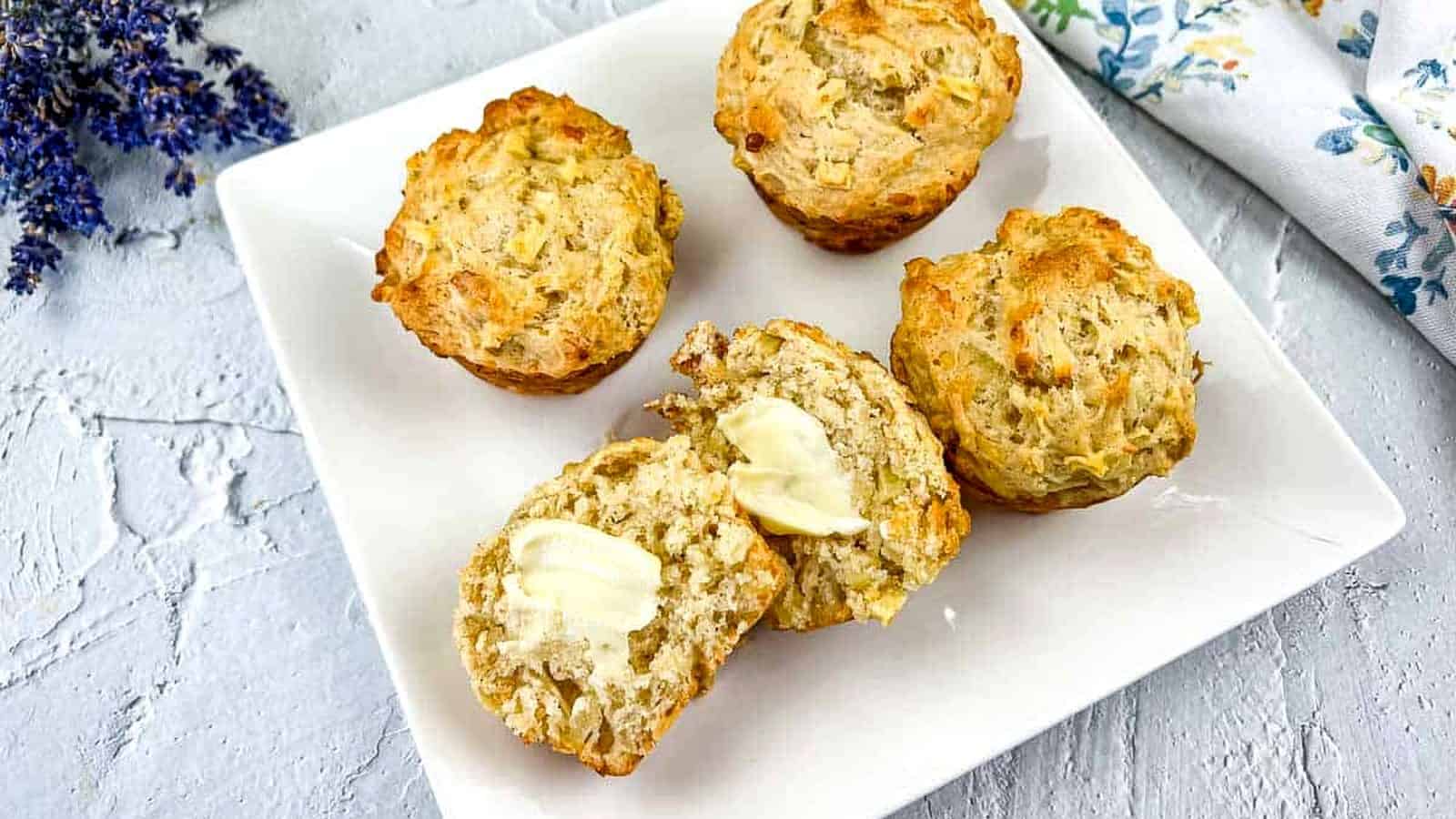 Apple cheddar muffins on a white plate, with one muffin cut open and spread with butter, showing the moist, cheesy interior.