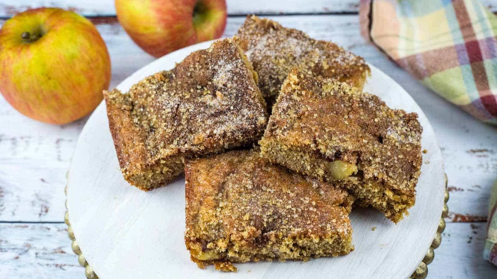 Four pieces of apple cider donut blondies are arranged on a white plate, with two whole apples and a plaid napkin in the background on a wooden surface.