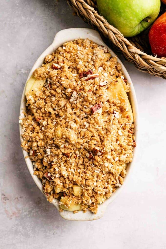 Oval baking dish filled with unbaked apple crumble topped with oat and nut streusel, next to a basket with green and red apples.