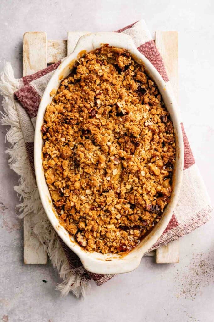 An oval baking dish filled with golden brown fruit crumble, placed on a red and white towel over a wooden board.