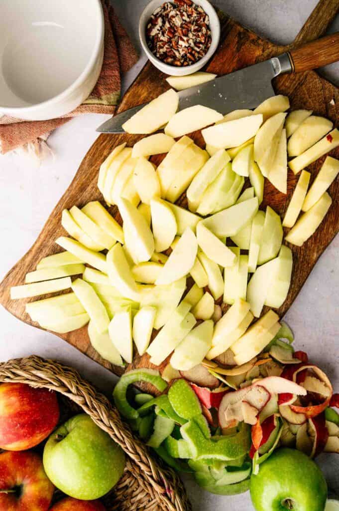 Sliced apples on a wooden cutting board with a knife, a small bowl of chopped nuts, apple peels, a napkin, and a basket of red and green apples nearby.
