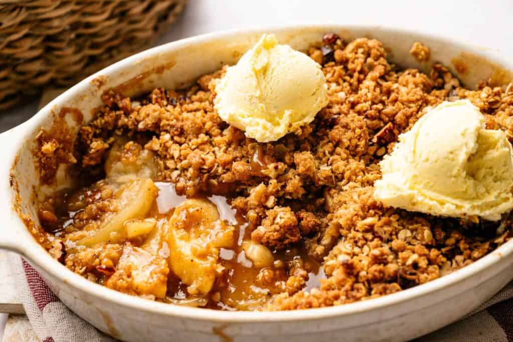 Baked apple crisp topped with a golden oat crumble and two scoops of vanilla ice cream in a white dish.