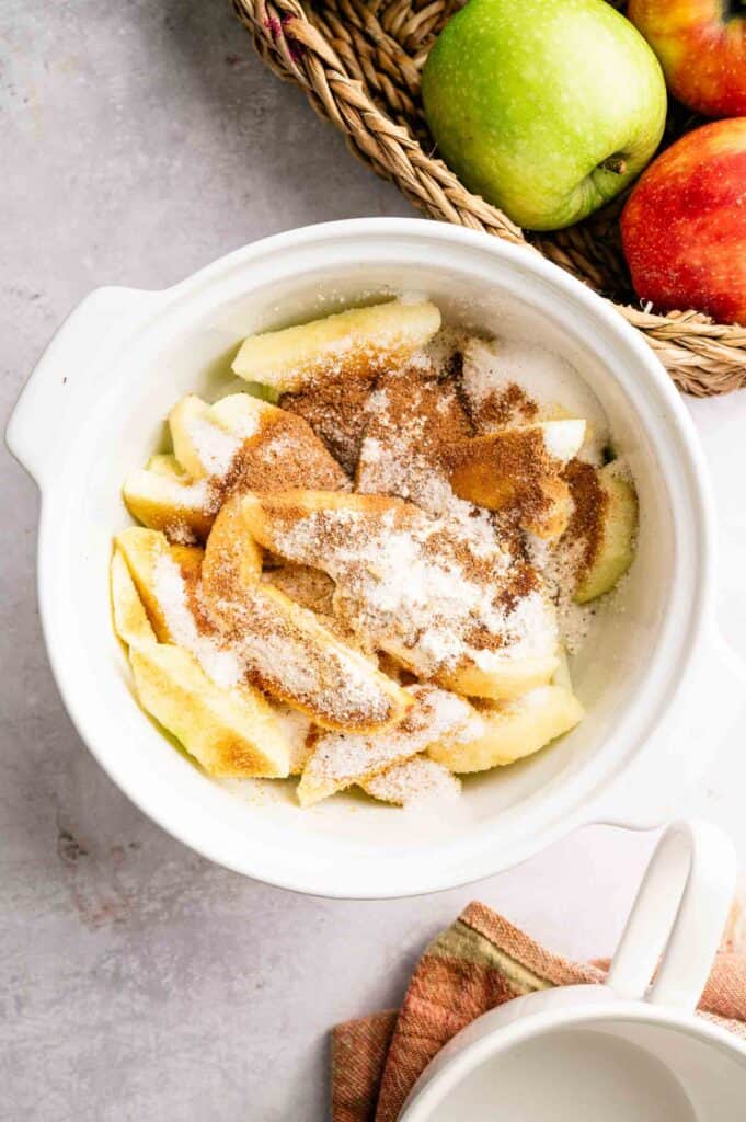 A white bowl filled with sliced apples topped with sugar and cinnamon, next to a basket containing green and red apples.