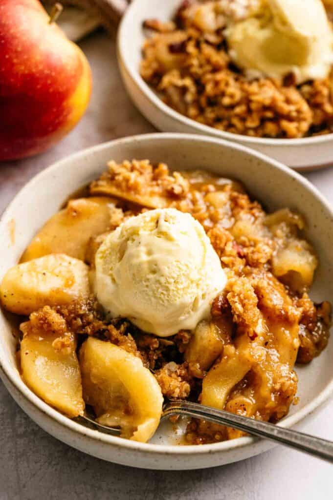 A bowl of apple crisp topped with a scoop of vanilla ice cream, with another bowl and an apple in the background.