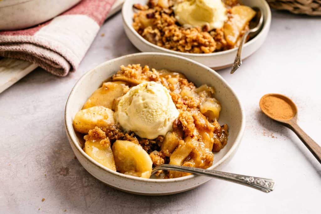 A bowl of apple crisp topped with a scoop of vanilla ice cream, with a spoon in the bowl and a small dish of ground cinnamon nearby.