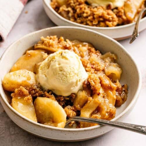 Bowl of apple crisp topped with a scoop of vanilla ice cream, with a spoon, placed on a light-colored surface. Another bowl and a wooden spoon with cinnamon are in the background.