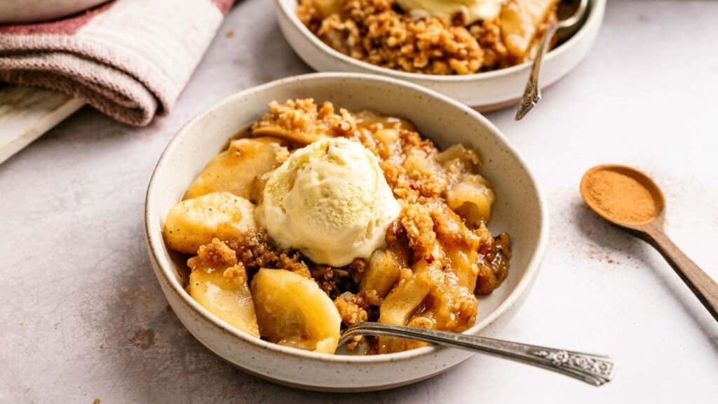 A bowl of apple crisp topped with a scoop of vanilla ice cream sits on a table, with a spoon inside and a wooden spoon with cinnamon nearby.