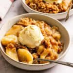 A bowl of apple crisp topped with a scoop of vanilla ice cream sits on a table, with a spoon inside and a wooden spoon with cinnamon nearby.