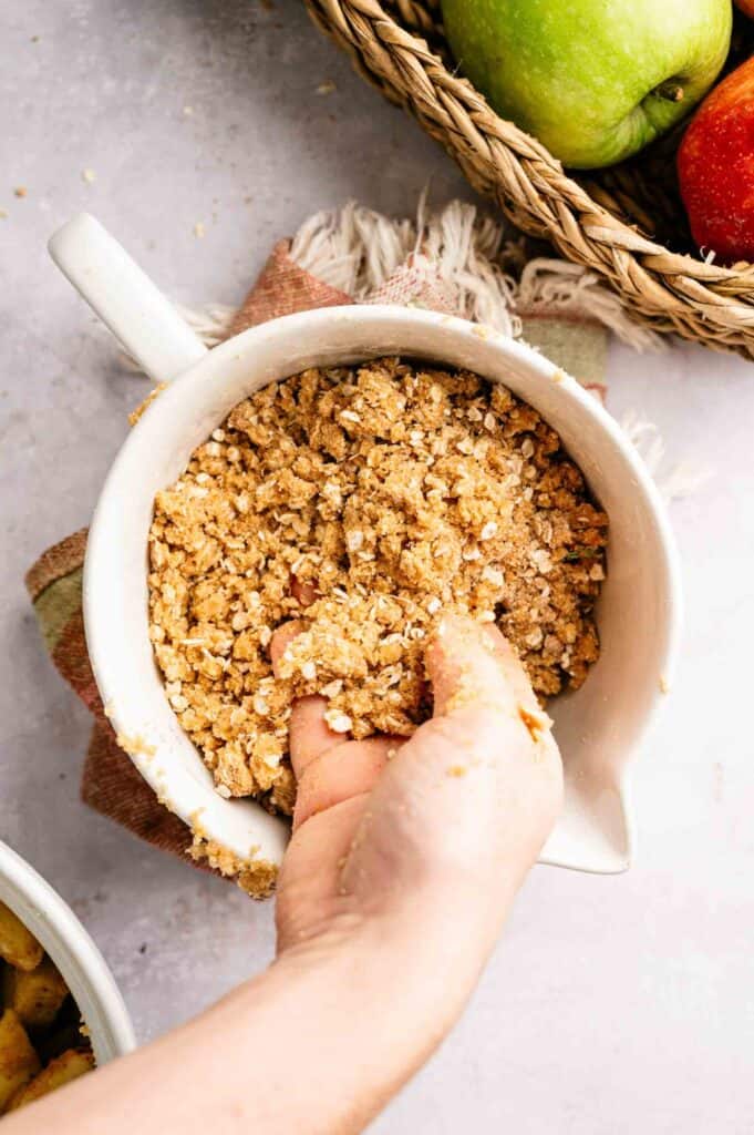 A hand mixes crumbly oat topping in a white bowl, with a basket of apples and a textured cloth visible nearby on a light surface.