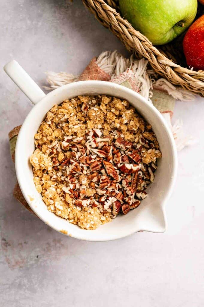 A white mixing bowl filled with oat and pecan crumble topping sits next to a basket holding a green apple and a red apple.
