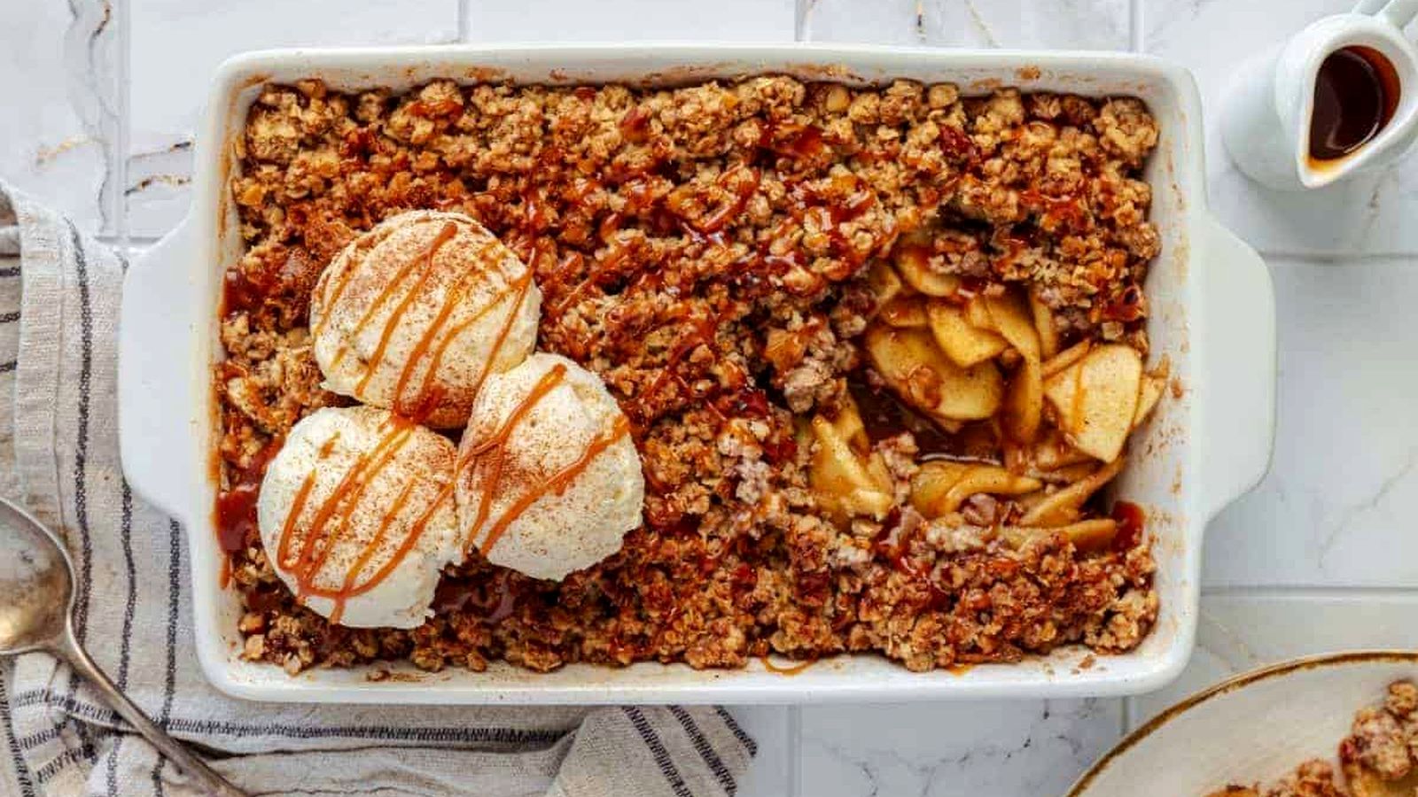 An Apple Crisp with Vanilla Ice Cream, served in a rectangular dish, topped with two scoops of vanilla ice cream and a caramel drizzle, accompanied by a portion on a plate beside it.