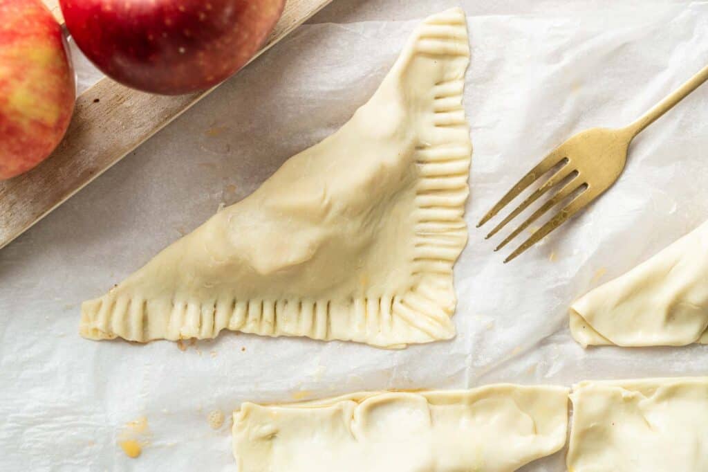 Unbaked apple turnovers on parchment paper next to a gold fork and fresh apples.