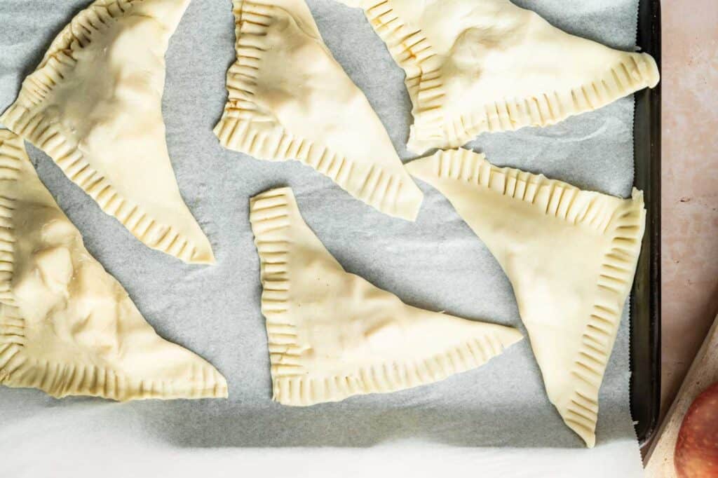 Unbaked triangular pastry turnovers arranged on a parchment-lined baking sheet, with crimped edges visible.