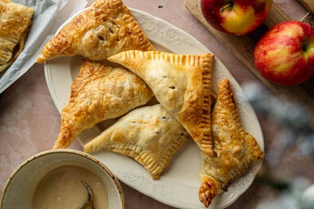 Four golden-brown, triangular apple turnovers on a white oval plate, with fresh apples and a bowl of glaze nearby on a light-colored surface.