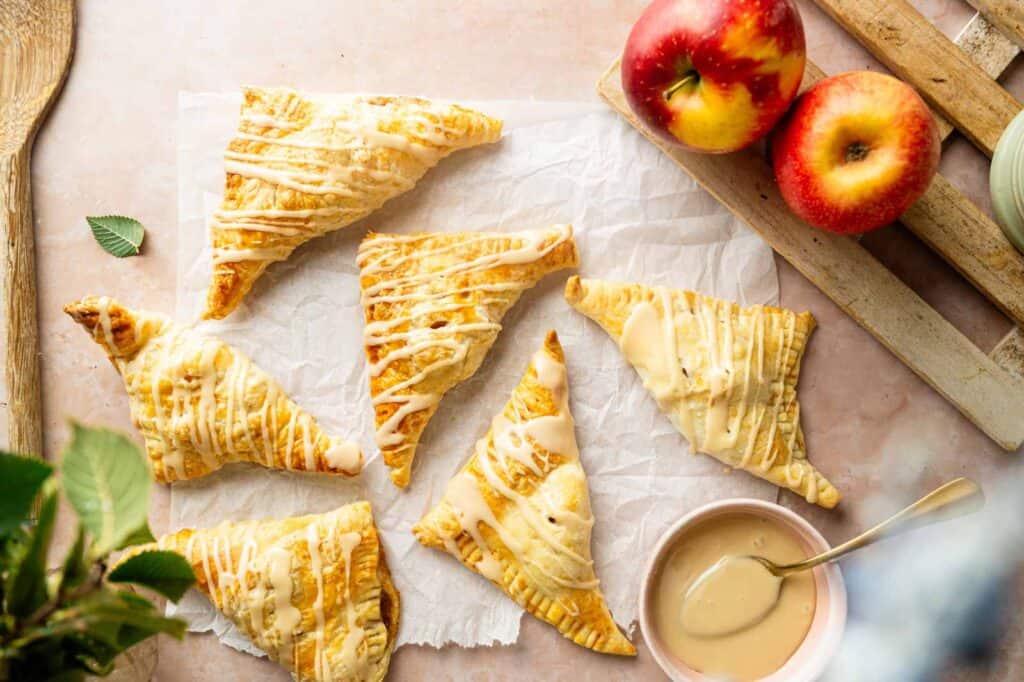 Five glazed apple turnovers on parchment paper, with two whole apples, a bowl of glaze with a spoon, and a wooden tray visible on a light surface.