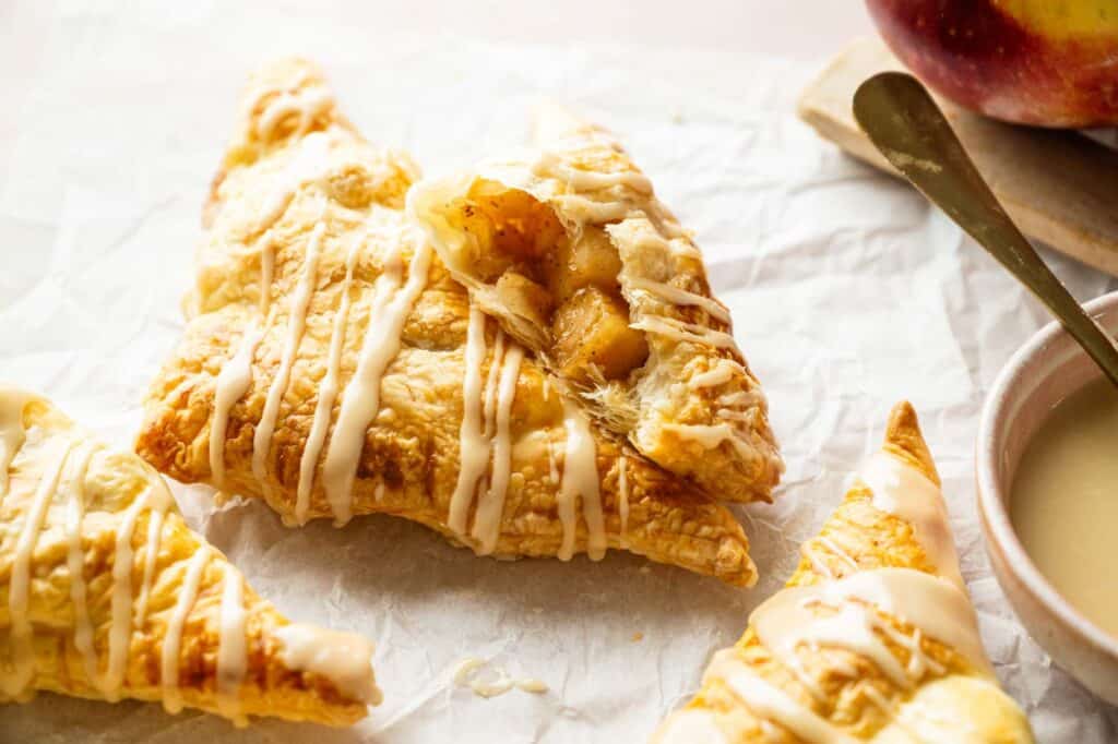 A close-up of glazed apple turnovers on parchment paper, with one turnover partially broken open to reveal the apple filling inside.