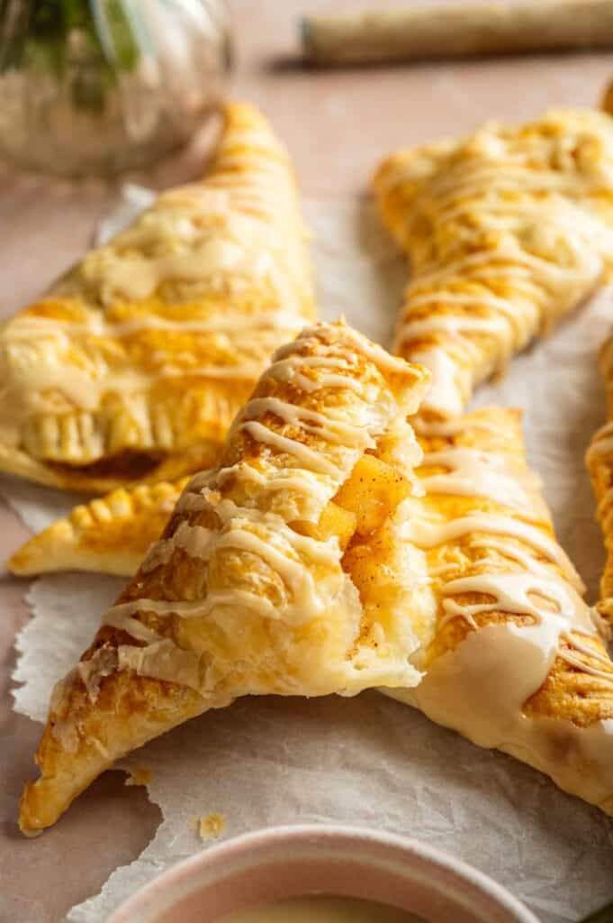 Golden apple turnovers with a flaky crust, drizzled with icing, displayed on parchment paper, with one turnover broken open to show the apple filling.