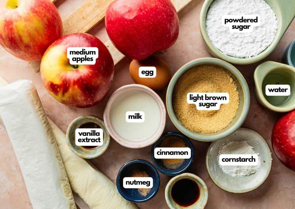 Assorted ingredients on a counter include apples, puff pastry, eggs, milk, vanilla, nutmeg, cinnamon, light brown sugar, powdered sugar, cornstarch, and water.