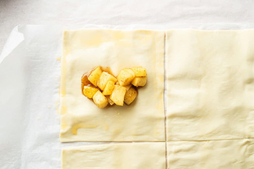 Four squares of pastry dough on parchment paper, with a small pile of diced, seasoned apples placed in the center of the top left square.