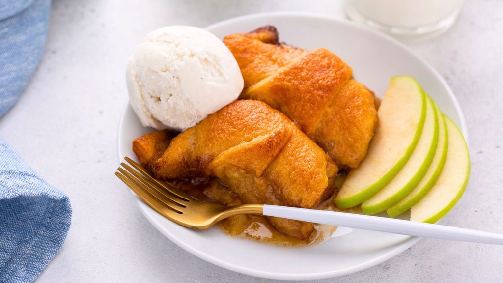 A plate with two baked crescent rolls, a scoop of vanilla ice cream, green apple slices, and a gold fork&mdash;perfect for celebrating National Apple Dumpling Day.