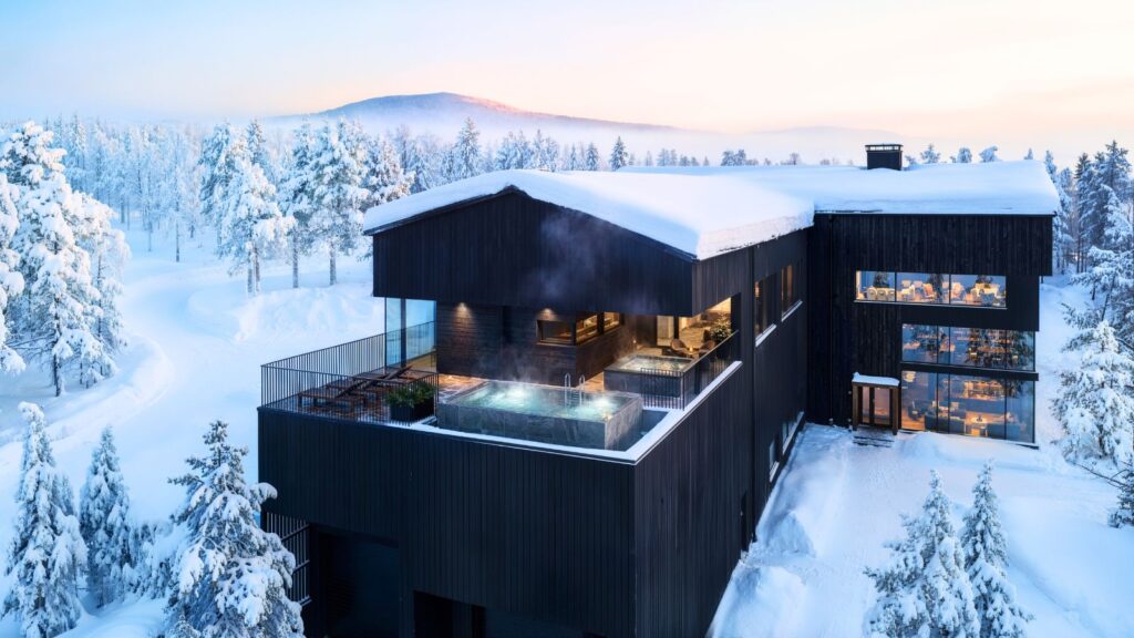 Large black building with outdoor hot tubs and steam rising, surrounded by snow-covered trees, during winter at sunset; mountains visible in the distance.