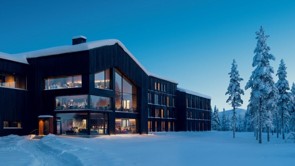Modern, dark-wood building with large windows sits in a snowy landscape surrounded by snow-covered trees at dusk.