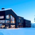 Modern, dark-wood building with large windows sits in a snowy landscape surrounded by snow-covered trees at dusk.